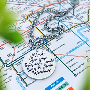 Op de afbeelding: Een zilverkleurige ketting met een ronde hanger met de tekst "Mind the gap between your life & your dreams". De ketting ligt op een Londense metrokaart, met groene bladeren op de voorgrond.