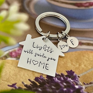 Può includere: Un portachiavi color argento con un ciondolo a forma di casa che recita "Lights will guide you HOME" in caratteri neri. Include due ciondoli rotondi più piccoli con le lettere "I ❤️ W" e "F". Il portachiavi è su un anello d'argento.