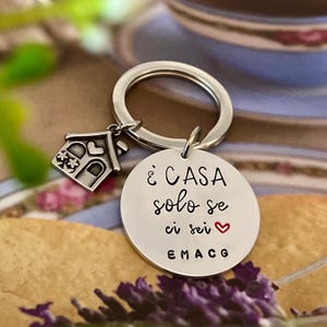 Puede incluir: Un llavero plateado con un colgante redondo y un charm de casa. El colgante tiene el texto "&Egrave; CASA solo se ci sei" con un coraz&oacute;n rojo. El charm de la casa tiene una puerta y ventanas en forma de coraz&oacute;n.
