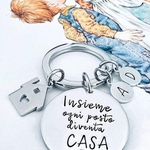 Puede incluir: Un llavero plateado con un dije en forma de casa, un dije redondo con texto en italiano y dijes de letras. El dije redondo dice "Insieme ogni posto diventa CASA" con un s&iacute;mbolo de coraz&oacute;n. Una ilustraci&oacute;n en acuarela de dos ni&ntilde;os est&aacute; en el fondo.