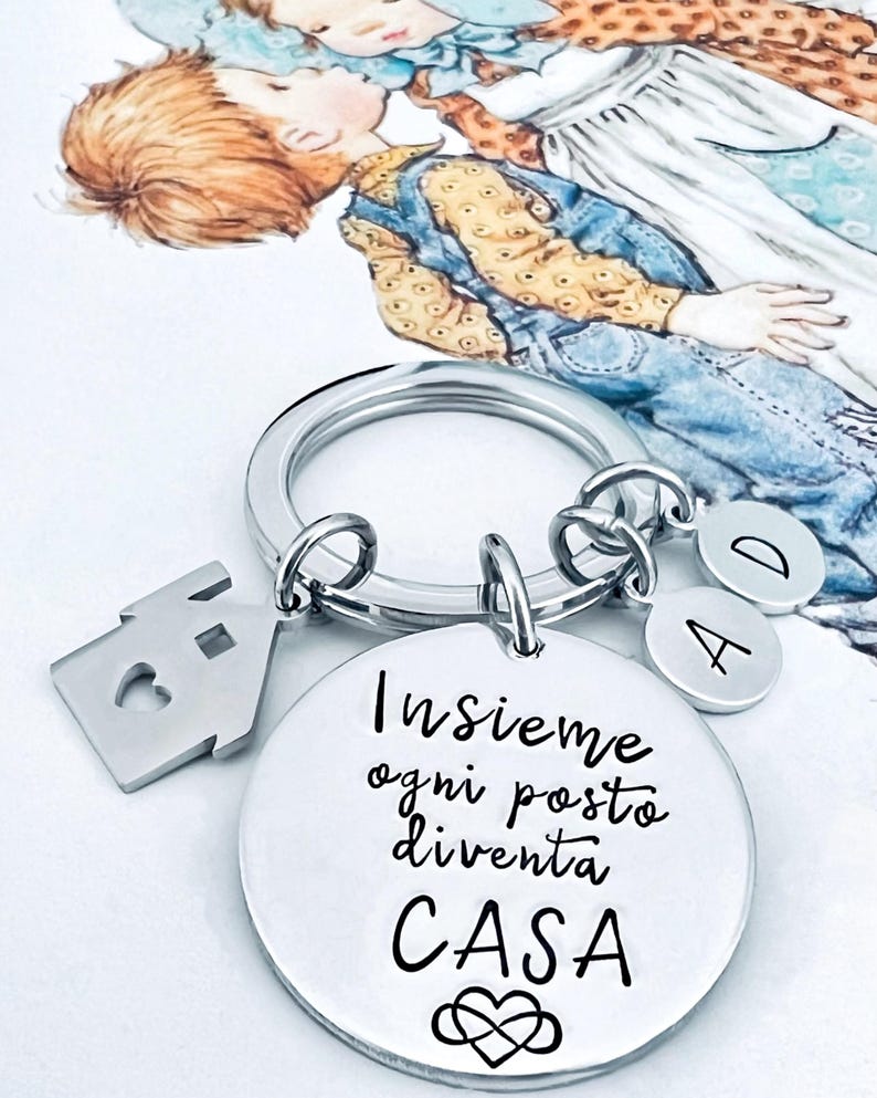 Puede incluir: Un llavero plateado con un dije en forma de casa, un dije redondo con texto en italiano y dijes de letras. El dije redondo dice "Insieme ogni posto diventa CASA" con un s&iacute;mbolo de coraz&oacute;n. Una ilustraci&oacute;n en acuarela de dos ni&ntilde;os est&aacute; en el fondo.