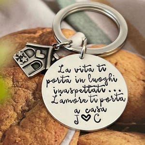 Puede incluir: Un llavero plateado con un colgante redondo y un charm de casa. El colgante tiene el texto italiano "La vita ti porta in luoghi inaspettati... L'amore ti porta a casa J ♡ C". El charm de casa tiene una puerta y detalles florales.