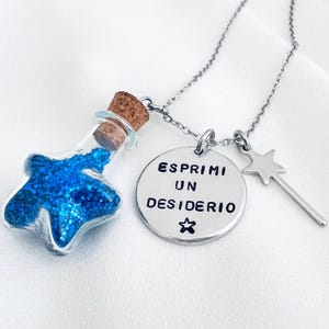 Puede incluir: Un collar de plata con una botella en forma de estrella llena de purpurina azul, un dije redondo con el texto "ESPRIMI UN DESIDERIO" y un dije de varita de estrella plateada.