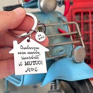 Può includere: Un portachiavi color argento con un ciondolo a forma di casa e un piccolo ciondolo rotondo. Il ciondolo a forma di casa ha un testo in italiano. Il ciondolo rotondo dice "TI AMO".