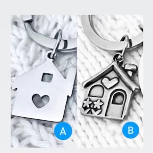 Puede incluir: Dos llaveros plateados con forma de casa. El llavero A tiene un recorte en forma de coraz&oacute;n y una ventana cuadrada. El llavero B presenta un coraz&oacute;n, flores, una chimenea y una puerta. Ambos llaveros est&aacute;n unidos a llaveros plateados.