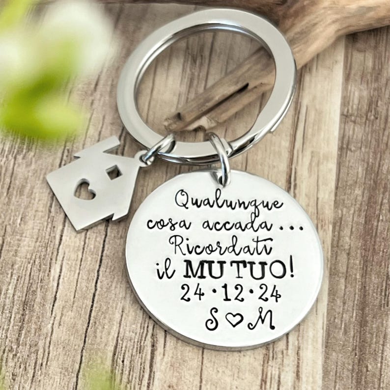 Puede incluir: Un llavero plateado con un colgante redondo y un charm en forma de casa. El colgante redondo tiene grabado el texto italiano "Qualunque cosa accada... Ricordati il MUTUO! 24&bull;12&bull;24 S♡M". El charm de la casa tiene un coraz&oacute;n recortado.