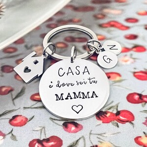 Puede incluir: Un llavero plateado con un dije en forma de casa y dos dijes con iniciales. El dije principal es un disco redondo con el texto italiano "CASA &egrave; dove sei tu MAMMA" y un s&iacute;mbolo de coraz&oacute;n. El llavero est&aacute; sobre un fondo floral.