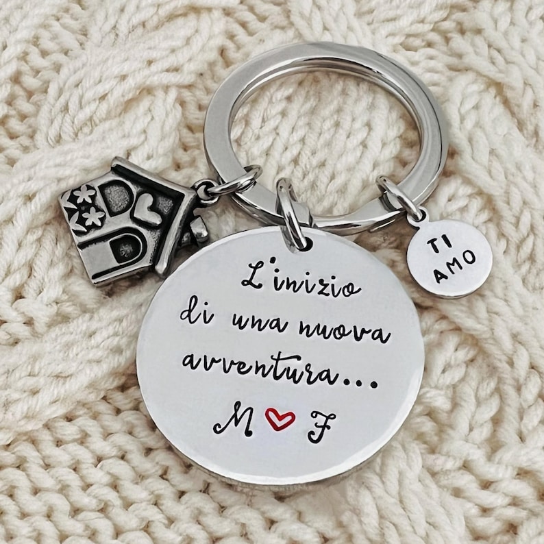 Puede incluir: Un llavero plateado con un dije de casa, un dije redondo con "TI AMO" y un dije redondo m&aacute;s grande con texto italiano y un coraz&oacute;n rojo. El llavero est&aacute; sobre una superficie de punto color crema.