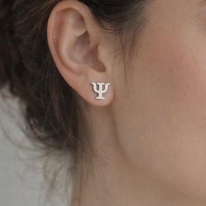 Può includere: Un orecchino a lobo color argento a forma di lettera greca Psi (Ψ) è mostrato su un orecchio. L'orecchino ha un design minimalista. Il simbolo Psi è spesso associato alla psicologia. L'orecchino è piccolo e semplice.