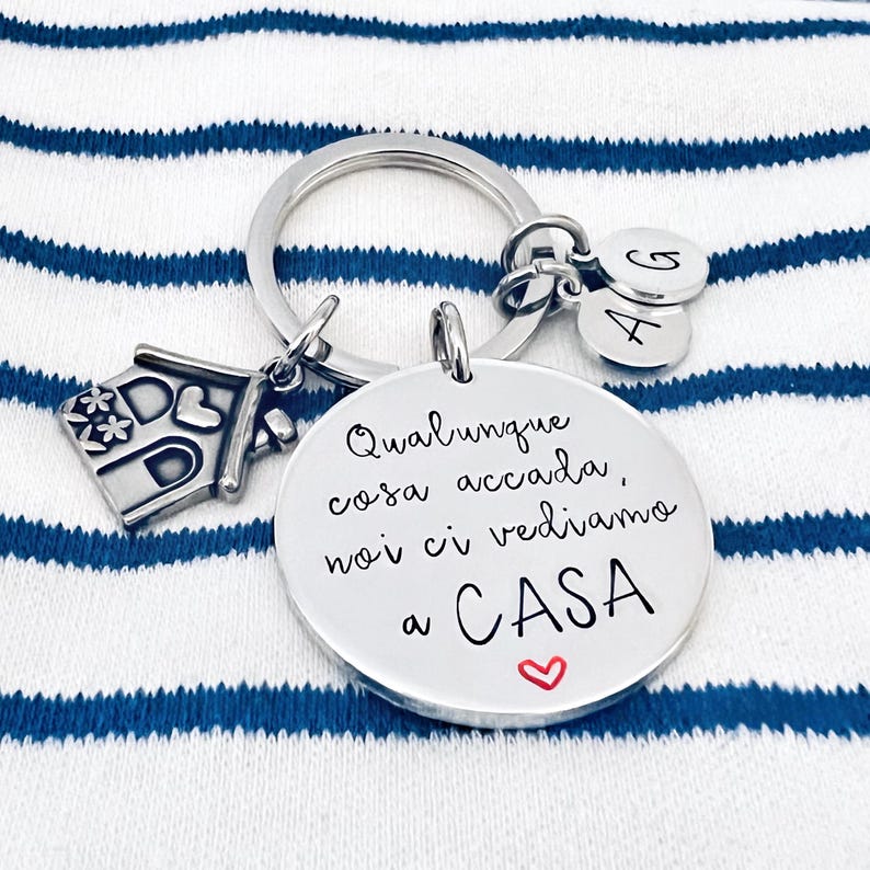 Puede incluir: Un llavero plateado con un dije de casa, dos dijes con letras y un dije redondo con texto en italiano y un coraz&oacute;n. El texto dice "Qualunque cosa accada, noi ci vediamo a CASA."
