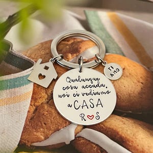 Puede incluir: Un llavero plateado con tres colgantes. Un colgante es una casa con un coraz&oacute;n recortado, otro es un colgante redondo con las palabras "TI AMO", y el tercero es un colgante redondo con texto italiano. El llavero est&aacute; sobre un fondo borroso.