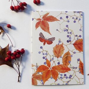 Op de afbeelding: Een wit notitieboek met een kleurrijk herfstbladerenontwerp. De omslag heeft een vlinder met rode en zwarte vleugels, oranje en bruine bladeren en paarse bessen.