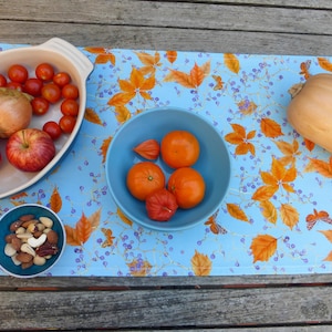 Puede incluir: Un camino de mesa azul con un patrón de hojas de otoño presenta una variedad de productos frescos, incluyendo pimientos rojos, tomates, manzanas, cebollas, calabaza butternut y naranjas. También se incluye un pequeño tazón de nueces.