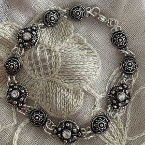Può includere: Bracciale color argento con elementi decorativi alternati. Il bracciale presenta accenti circolari con perline e design rotondi traforati con pietre centrali chiare e scintillanti. Il bracciale è collegato con piccole catene d'argento ed è esposto su una superficie strutturata di colore neutro.