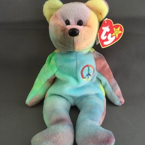 May include: A colorful plush bear with a peace sign on its chest. The bear has a tie-dye pattern in shades of blue, green, pink, yellow, and purple. It has a red heart-shaped tag attached to its ear.