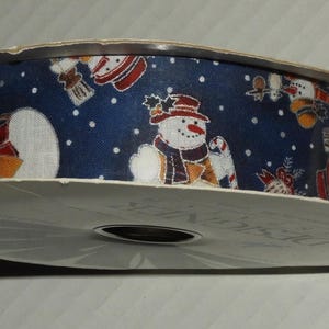 May include: A roll of Christmas ribbon with a navy blue background and a repeating pattern of snowmen, snowflakes, and other holiday designs. The ribbon is on a white spool.