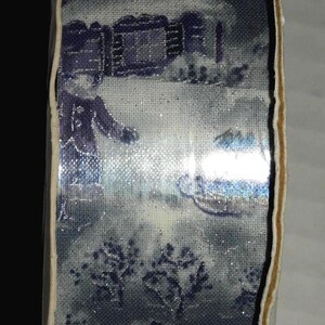 May include: A roll of decorative ribbon featuring a winter scene with a house, trees, and a person. The design is in shades of blue and white, with a glittery effect. The ribbon has a cream-colored edge.