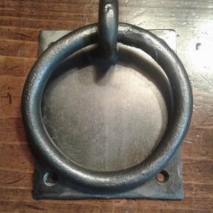 May include: A dark gray metal ring attached to a square metal plate. The ring is circular and has a thick, rounded profile. The plate has two holes for mounting. The metal has a slightly textured appearance.