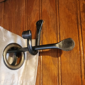 May include: A wrought iron curtain tieback with a dark finish. The tieback features a curved arm that holds a curtain ring and a decorative, flattened end. The hardware is mounted on a wooden surface.