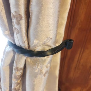 以下が含まれることがあります： 黒いねじれた金属製のカーテンタッセルが、花柄のベージュ色のカーテンを留めています。タッセルはカーテンに巻き付けられており、端が見えています。