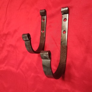 May include: Two handcrafted, dark metal wall hooks. Each hook has a curved, J-shaped design with a flat, rectangular mounting plate featuring three holes for screws. The hooks are displayed against a red background.