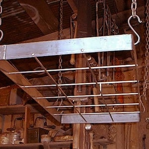 Puede incluir: Un estante rectangular de metal para ollas, suspendido del techo por cadenas y ganchos. El estante tiene una rejilla de barras horizontales para sostener ollas y sartenes. El fondo muestra un taller desordenado.