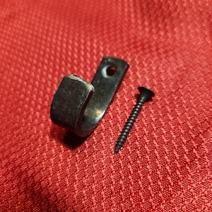 May include: A black metal hook with a curved end and a screw. The hook has a small hole for mounting. The screw is black and has a pointed tip. The items are on a red textured surface.