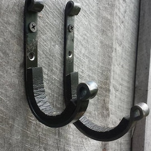 Puede incluir: Dos ganchos de metal negro montados sobre una superficie de madera envejecida. Cada gancho tiene un diseño curvo con un extremo redondeado y se fija con tornillos. Los ganchos son para colgar objetos.