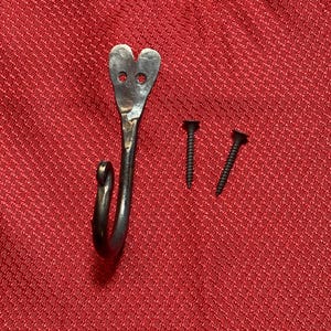 May include: Hand-forged metal hook with a heart-shaped top and two screw fasteners. The hook is dark gray and has a curved design. The screws are black. The hook is designed for hanging items.