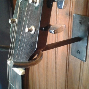 May include: Acoustic guitar headstock mounted on a custom metal wall hanger. The guitar's tuning pegs are visible, along with the strings and a portion of the fretboard. The hanger is made of dark metal and is attached to a wooden wall.