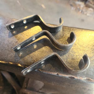 Puede incluir: Tres soportes artesanales de barra de cortina de metal oscuro. Cada soporte tiene un diseño curvo con un gancho en el extremo y agujeros para el montaje. Los soportes se muestran sobre una superficie metálica texturizada, mostrando su estilo rústico e industrial.