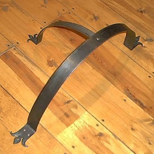 May include: Two curved, dark metal brackets with decorative fleur-de-lis ends. The brackets are positioned on a light-colored wooden surface, creating a contrast between the metal and the wood.