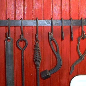 Op de afbeelding: Een smeedijzeren pottenrek met zeven hangende gereedschappen tegen een rode houten muur. De gereedschappen omvatten een platte spatel, een lusvormige haak, een spiraalvormig gereedschap, een gebogen gereedschap en een hartvormig gereedschap.