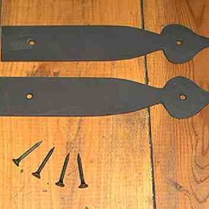 May include: Two black metal strap hinges with decorative ends and pre-drilled holes, plus several black screws. The hinges are on a wooden surface, showcasing a rustic hardware aesthetic.