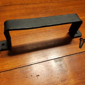 May include: A black, hand-forged metal handle with a rectangular design. The handle has a curved top and two mounting plates with pre-drilled holes. Two black screws are included.