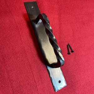 Peut inclure: Une poignée de porte en métal forgé à la main avec une poignée torsadée. La poignée est montée sur une plaque rectangulaire avec un bord martelé. Deux vis noires sont incluses pour l'installation. Le métal a une finition sombre et rustique.