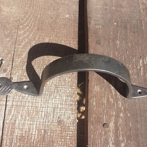Może przedstawiać: Kuty żelazny uchwyt do drzwi w kolorze ciemnoszarym o zakrzywionym kształcie i liściastych zakończeniach. W zestawie cztery srebrne śruby. Uchwyt jest pokazany na zwietrzałej drewnianej powierzchni.