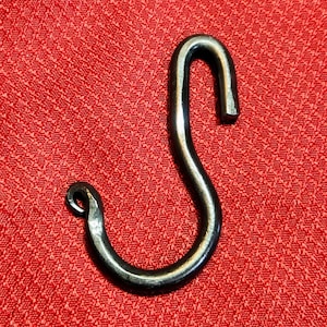 May include: Hand-forged, dark metal S-hook. The hook is curved and has a loop at one end and a straight end at the other. The hook is set against a red textured background.