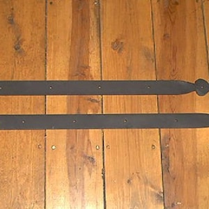 May include: Two black metal strap hinges with decorative heart-shaped ends. Each hinge has multiple pre-drilled holes for mounting. The hinges are displayed on a wooden surface with a warm, natural wood grain.