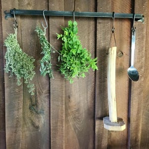 Puede incluir: Escena de cocina rústica con una barra colgante de metal oscuro. Manojos de hierbas secas, un portarrollos de papel de madera y una cuchara de metal cuelgan de la barra contra una pared de madera. Las hierbas son verdes y marrones.