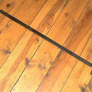 Może przedstawiać: Długa, ciemna metalowa korba z zagiętymi końcami, leżąca na drewnianej powierzchni. Uchwyt wydaje się być wykonany z litego metalu. Drewniana powierzchnia ma ciepłe, naturalne usłojenie.