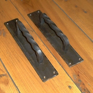 May include: Two black wrought iron door pulls with a twisted handle design. Each pull is mounted on a rectangular plate with rounded corners and screw holes. The door pulls are set against a light brown wooden surface.