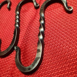 Peut inclure: Gros plan sur trois crochets en S en métal foncé forgés à la main. Les crochets ont un design torsadé sur la tige centrale et une extrémité incurvée. Le fond est un tissu rouge texturé.