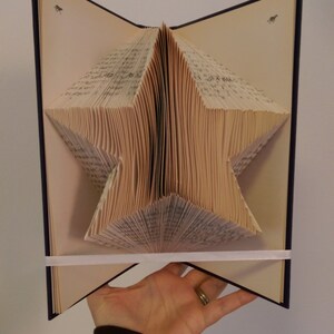 Può includere: Una scultura a forma di stella fatta a mano con le pagine di un libro. Il libro è aperto, con le pagine piegate per creare una forma di stella tridimensionale. Il libro ha una copertina blu scuro e un nastro bianco lo tiene aperto.