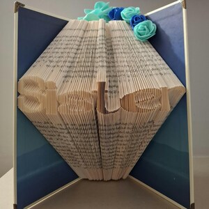 May include: A folded book art sculpture with the word Sister formed by the pages. The book is open with dark blue interior covers and is decorated with light blue and dark blue artificial foam roses at the top centre of the pages.