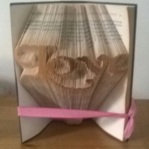 May include: A book sculpture with the word "Peace" carved into the pages. The book is held together with a pink ribbon. The pages are folded to create the letters, and the edges of the book are visible.