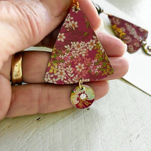 May include: Floral print earrings with a triangular top and a round bottom. The earrings feature a burgundy background with white and pink flowers and green leaves. A small, round charm with a clear crystal is attached to the bottom.