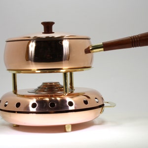 Peut inclure: Une fondue en cuivre avec une poignée en bois et un couvercle. La casserole repose sur une base ronde en cuivre avec des trous pour la circulation de la chaleur.