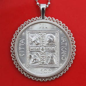 Könnte beinhalten: Silberne Halskette mit Anhänger in Form einer runden Münze mit einem dekorativen Rand. Die Münze zeigt ein zentrales Bild von vier Quadraten mit verschiedenen Symbolen und dem Text "Citta del Vaticano L.500".