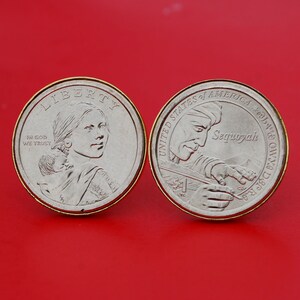 Op de afbeelding: Twee zilveren Amerikaanse dollarmunten met gouden randen. De voorzijde toont een portret van Sacajawea die haar baby op haar rug draagt. De achterzijde toont een portret van Sequoyah met de tekst "Sequoyah" en "United States of America" en het jaar "1999".